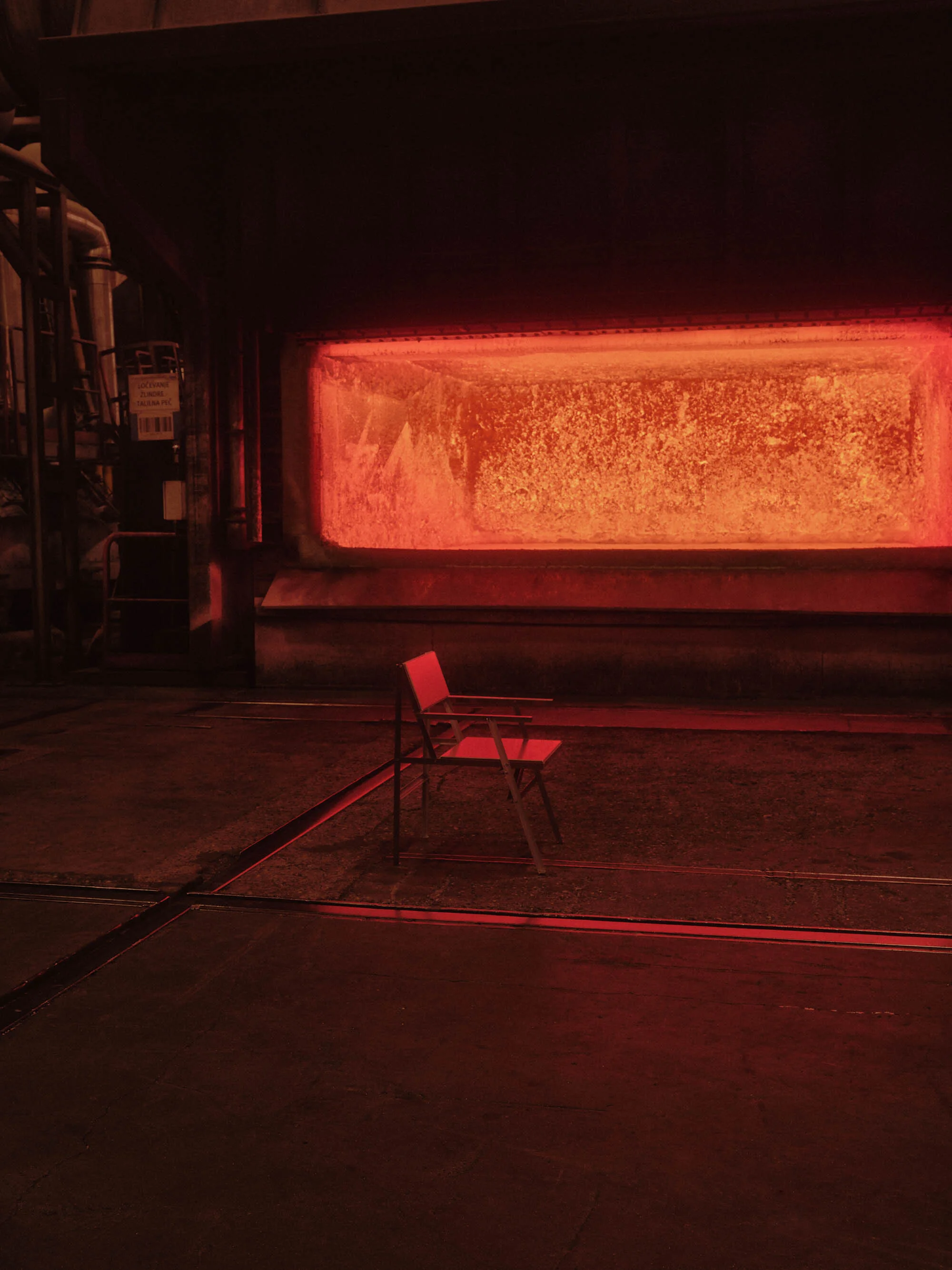 Metal foundry interior with a red metal folding chair in the foreground, against a backdrop of a fiery furnace and metal structures.