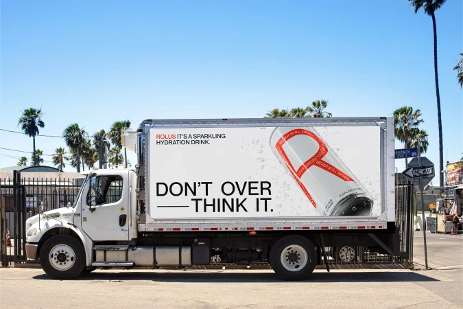 White box truck with a large advertisement for Rolus sparkling hydration drink, parked on a street with palm trees and blue sky.
