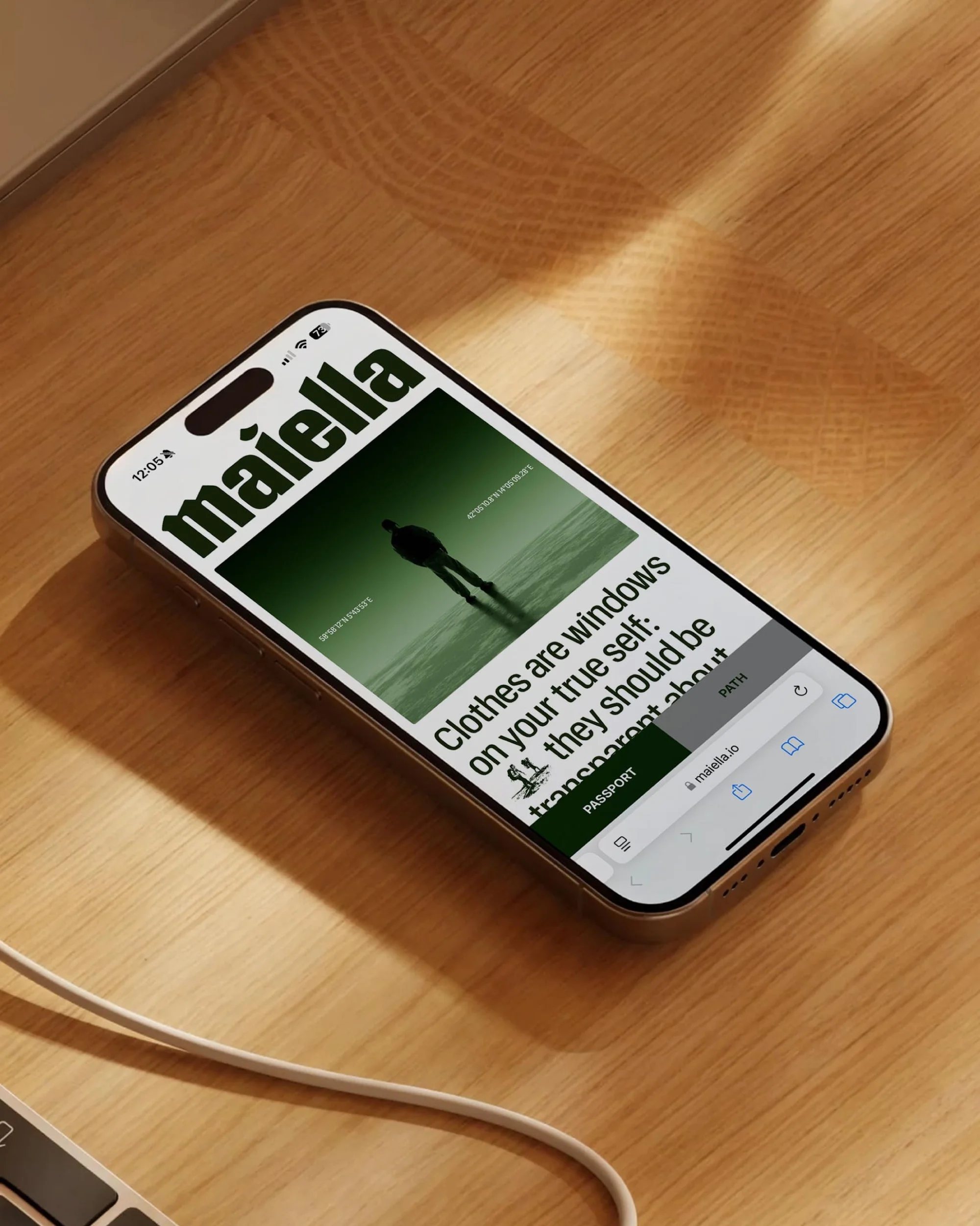 IPhone displaying Maiella website on a wood surface with a laptop corner and charging cable visible under soft studio lighting.