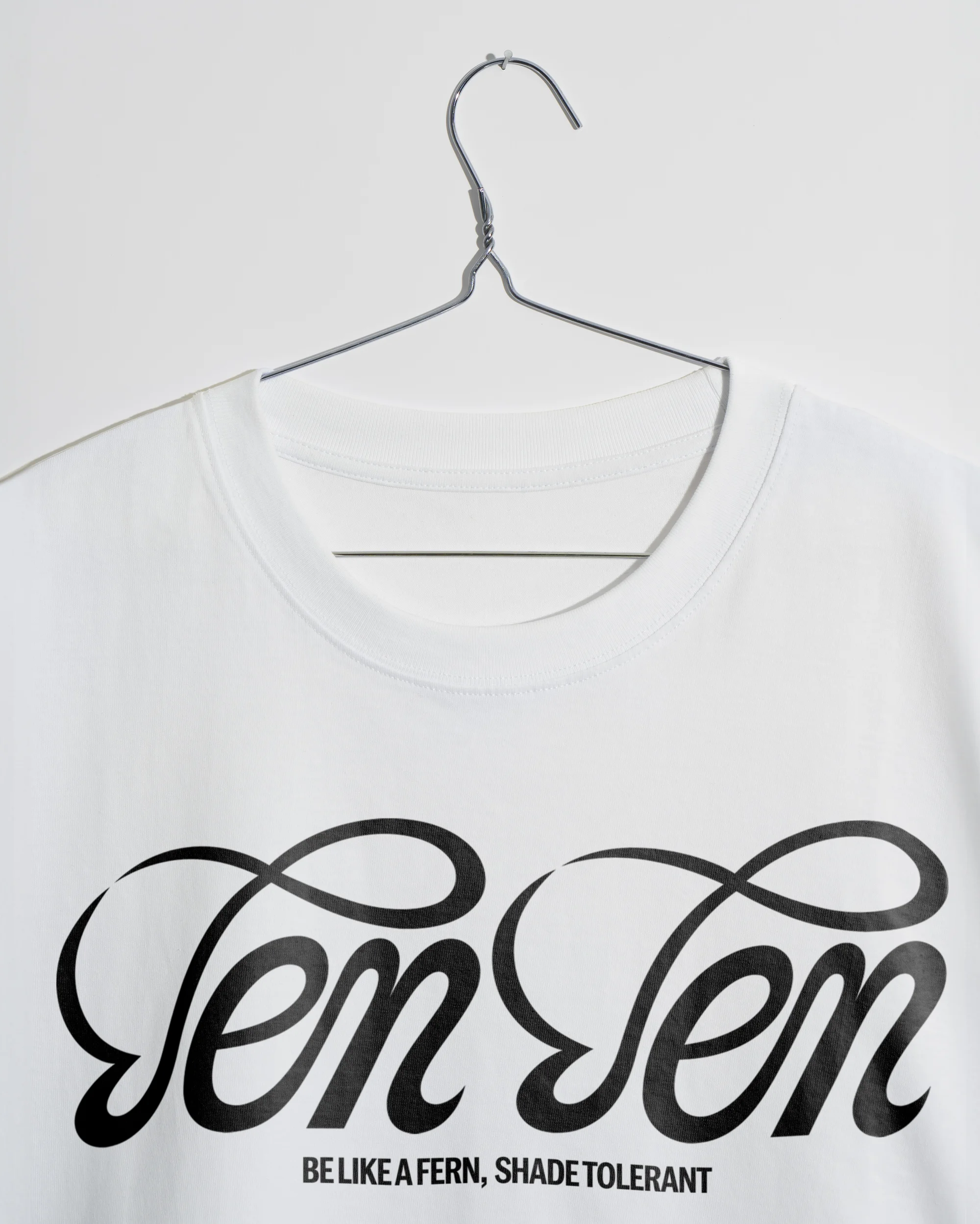 White t-shirt on a metal hanger against a white background with the text “Ten Ten” and “Be Like A Fern, Shade Tolerant” printed on the front.