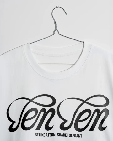 White t-shirt on a metal hanger against a white background with the text “Ten Ten” and “Be Like A Fern, Shade Tolerant” printed on the front.
