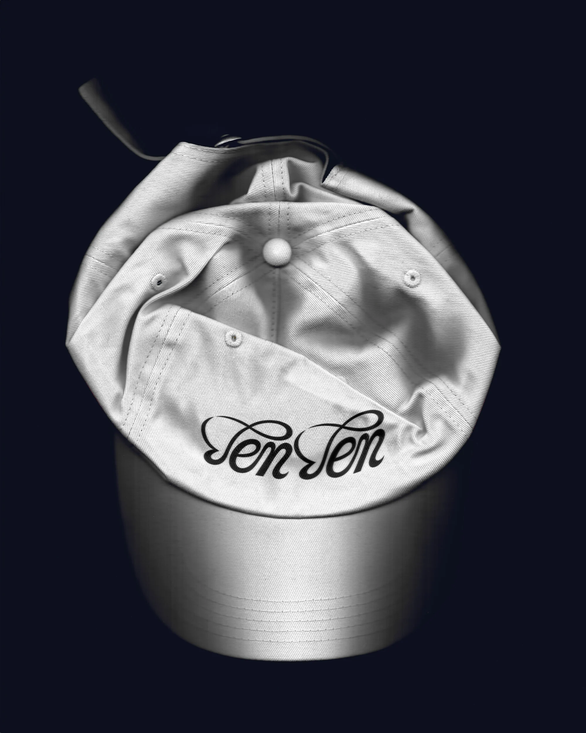 Upside-down white baseball cap with adjustable strap and embroidered 'Ten Ten' logo on a dark blue background in a studio setting.