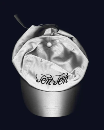 Upside-down white baseball cap with adjustable strap and embroidered 'Ten Ten' logo on a dark blue background in a studio setting.