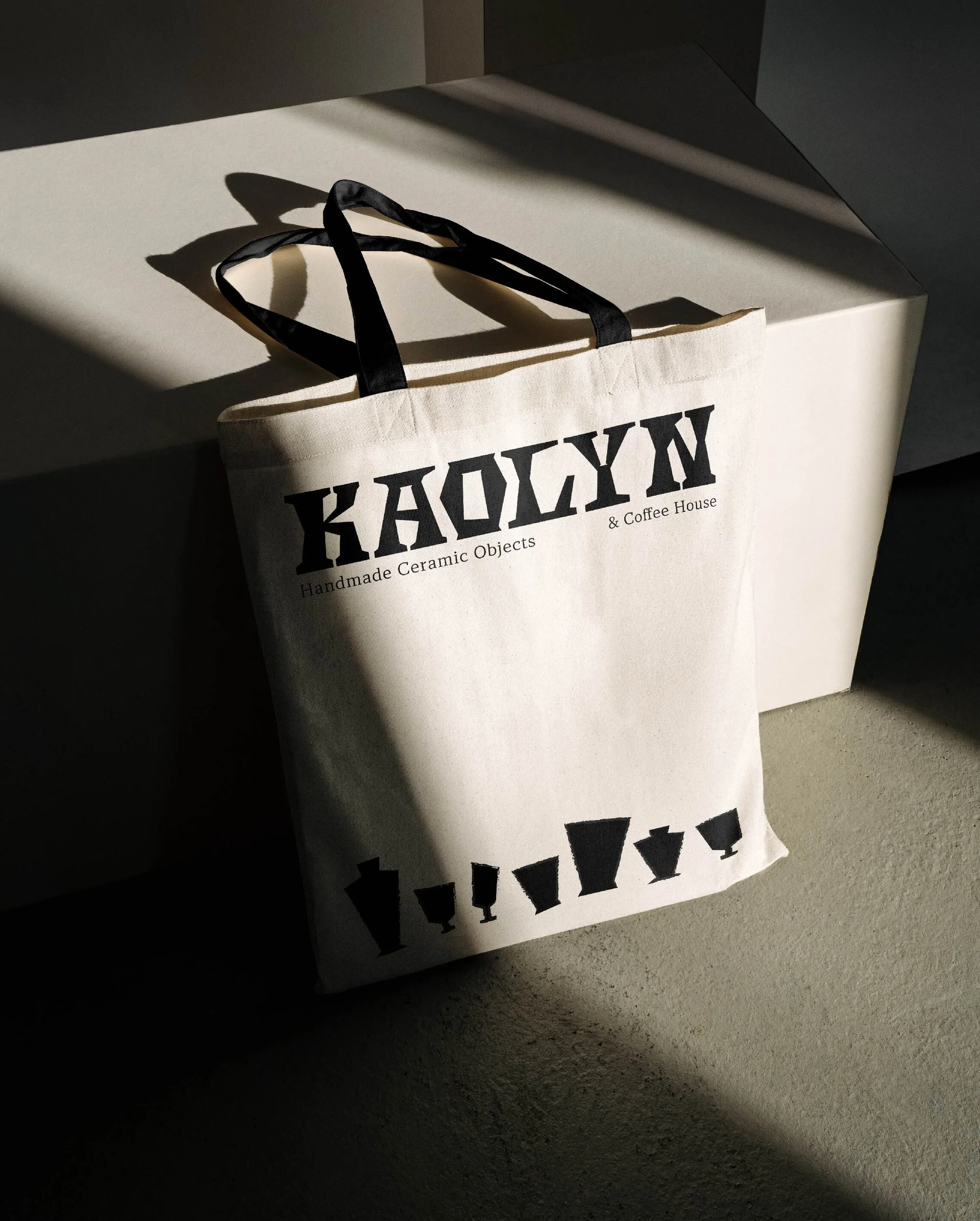 A beige tote bag with black handles sits upright in a brightly lit corner, featuring black text and graphics, with a cast shadow.