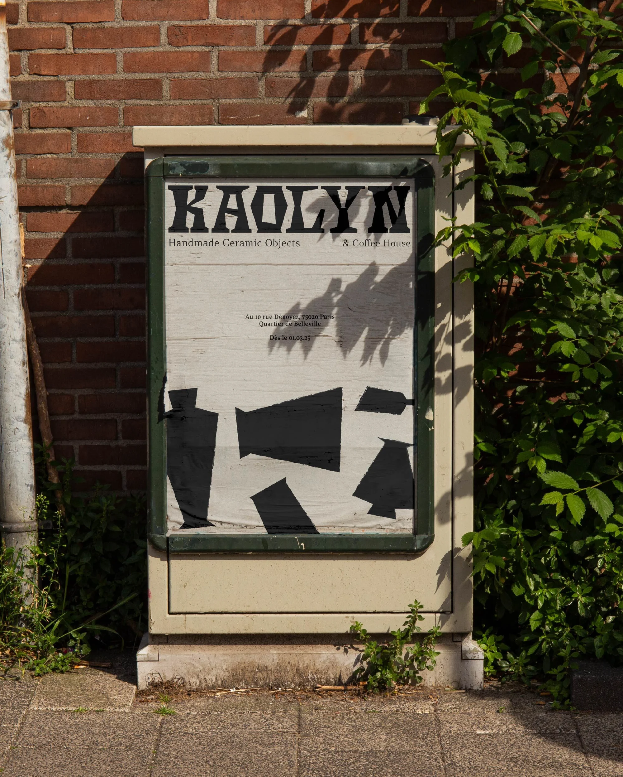 Outdoor poster display featuring a Kaolyn handmade ceramic object advertisement next to a red brick wall and greenery on a city sidewalk.