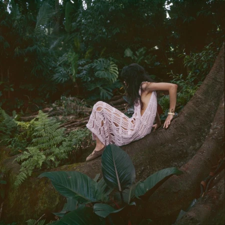 Woman in crochet dress sits on a tree trunk in a lush forest setting, surrounded by tropical plants and greenery.