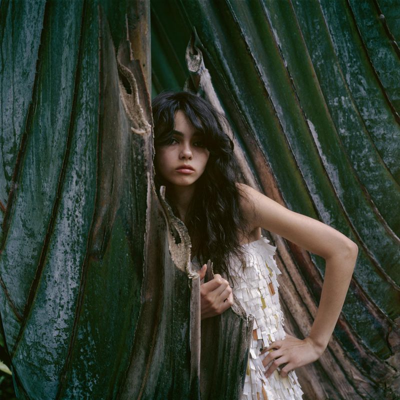 A young woman with long dark hair poses in a white dress with hanging accents, standing against a natural green tropical leaf backdrop.