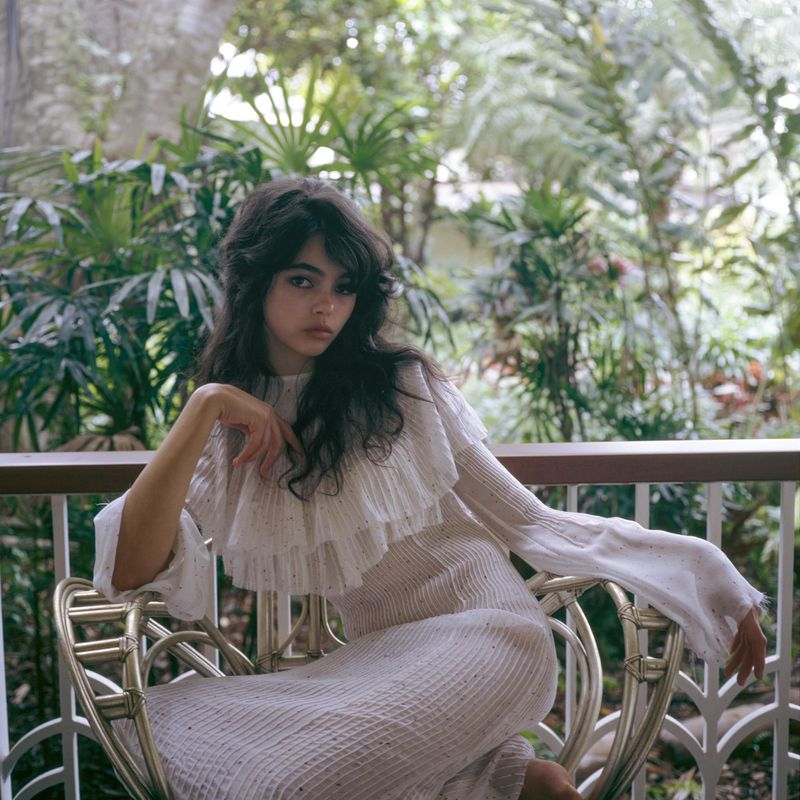 A young woman with dark wavy hair wearing a white dress is sitting on a rattan chair in front of a tropical green background.
