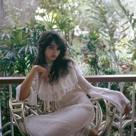 A young woman with dark wavy hair wearing a white dress is sitting on a rattan chair in front of a tropical green background.