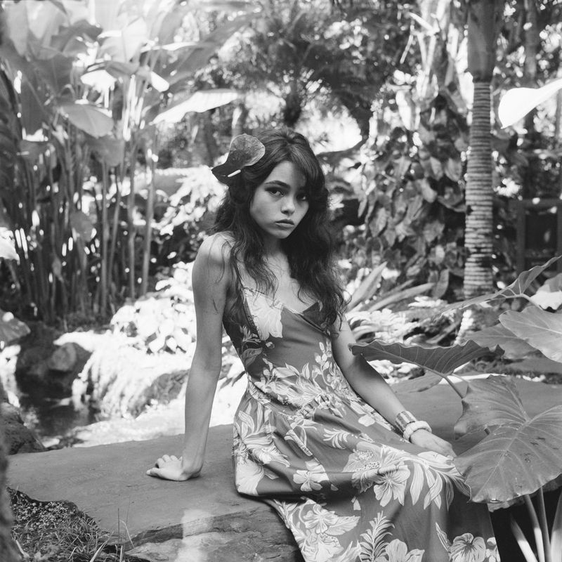 Black and white portrait of a woman in a floral dress, posing in a tropical garden setting surrounded by lush green foliage.