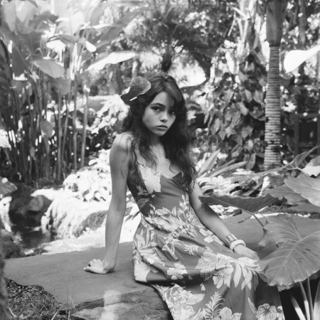 Black and white portrait of a woman in a floral dress, posing in a tropical garden setting surrounded by lush green foliage.