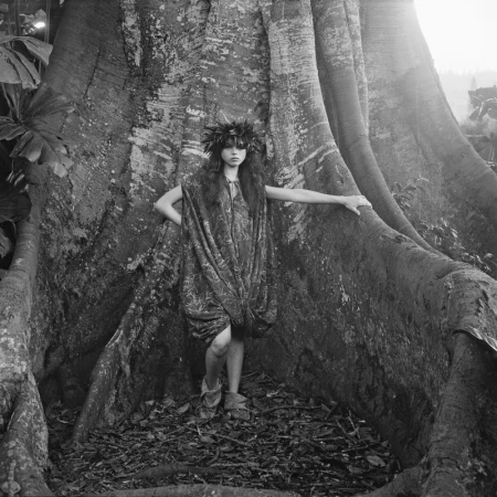 Woman in earth-toned dress and leaf crown poses at base of enormous banyan tree with large roots in dappled natural light.