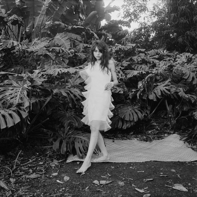 Young woman in a white ruffled dress poses barefoot in a tropical outdoor setting with large leafy green plants in a black and white photo.