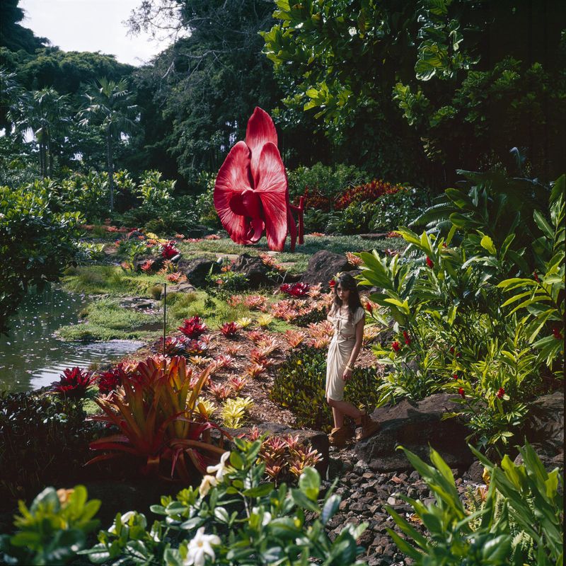 A woman in a light dress stands in a lush garden with a pond, greenery, and a large red botanical sculpture under natural lighting.
