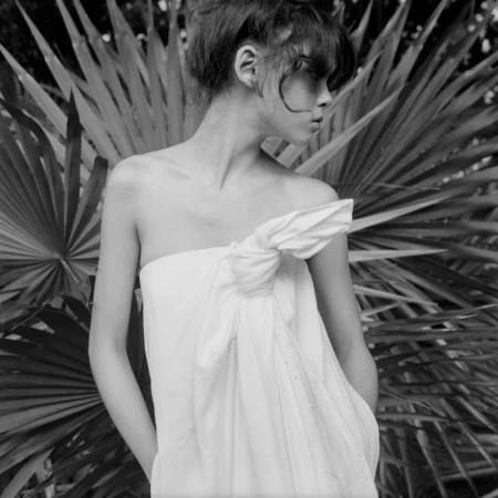 Monochrome portrait of a woman in a tied white garment, hands in pockets, against a backdrop of large palm fronds in soft light.