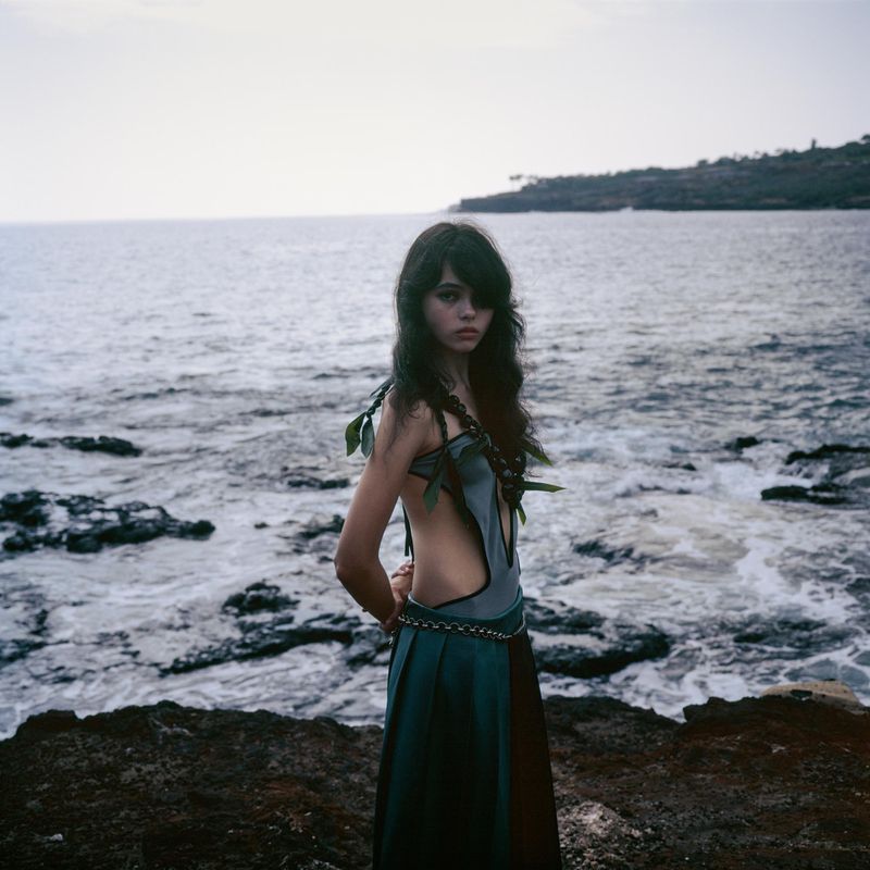 A woman stands on a rocky shore near the ocean, wearing a teal and brown dress with leafy accents and braids in her dark hair.