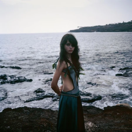 A woman stands on a rocky shore near the ocean, wearing a teal and brown dress with leafy accents and braids in her dark hair.