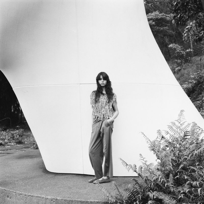 Young woman poses in loose pants and top against a large, curved white architectural structure in a monochrome outdoor fashion shot.