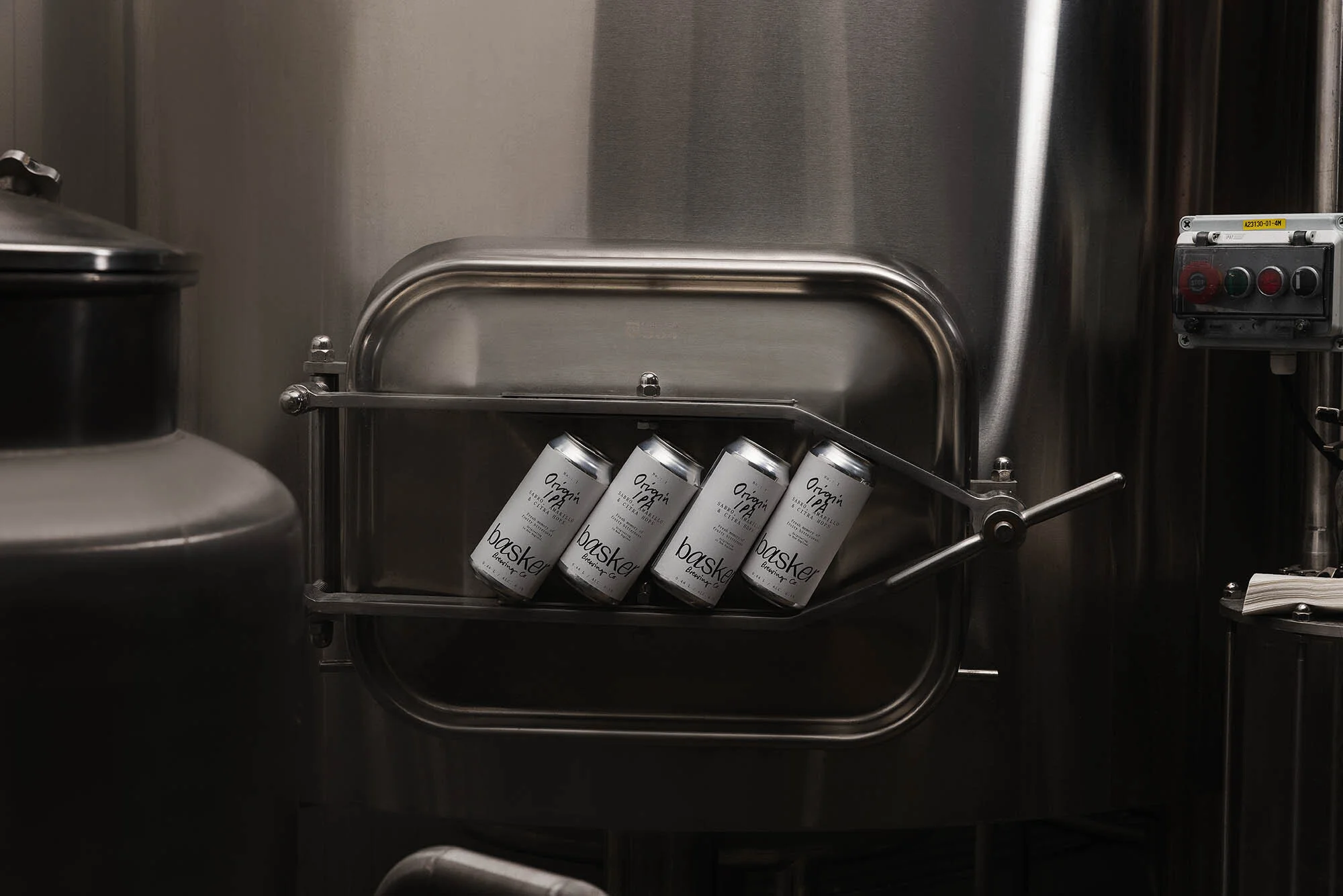 Four silver Basker Brewing Co. beer cans are placed in a shiny stainless-steel tank compartment, inside a brewery setting, in a medium shot.