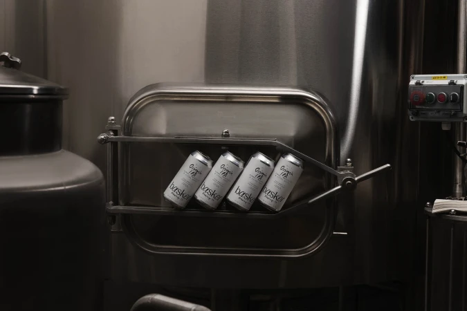 Four silver Basker Brewing Co. beer cans are placed in a shiny stainless-steel tank compartment, inside a brewery setting, in a medium shot.