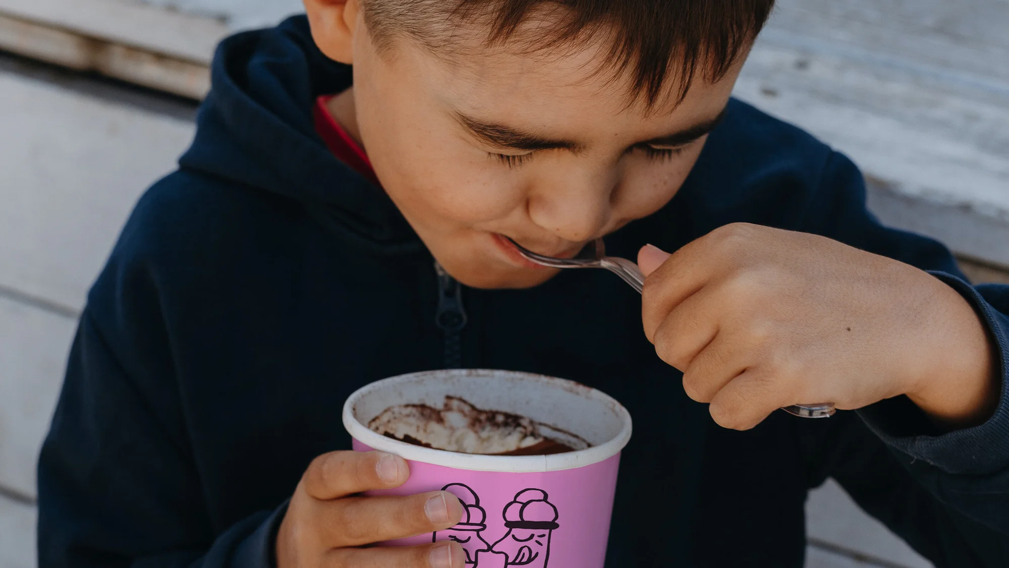 Boy eating chocolate ice cream from a pink cup with a spoon. Close-up shot of a child in a navy blue hooded sweatshirt outdoors.