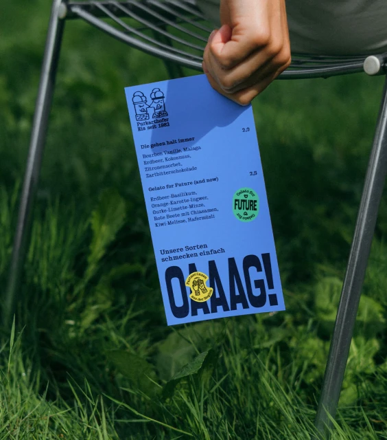 Hand holds blue menu card for 'OAAAG!' ice cream, listing flavors and prices, in an outdoor setting with green grass visible.