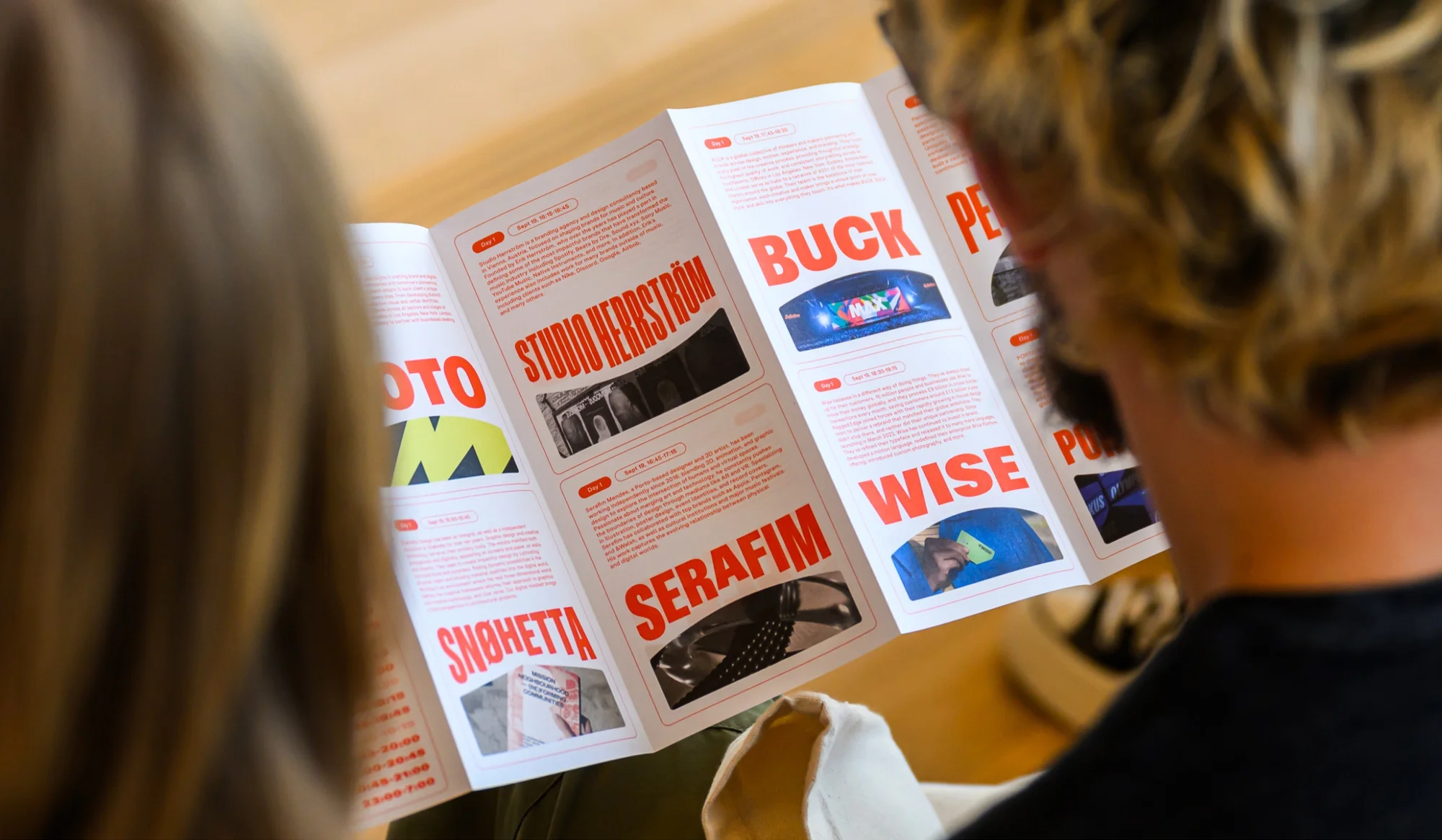 Two people view a brochure with typography, project photos, and event descriptions in a naturally lit room with a light wood floor.