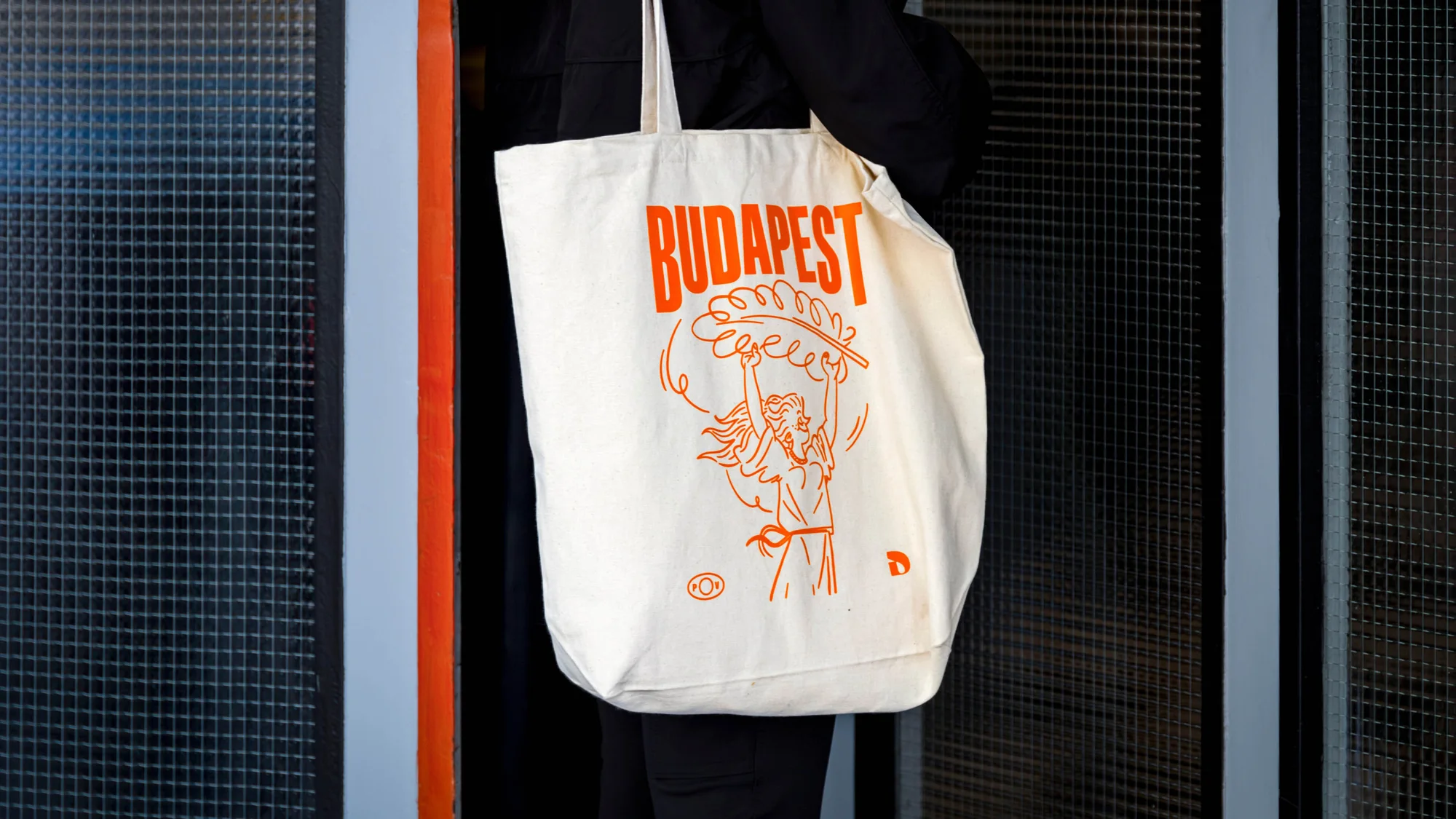 Cotton tote bag with orange Budapest illustration and a woman holding a plant. Worn by a person in a dark coat against a grey tile wall.