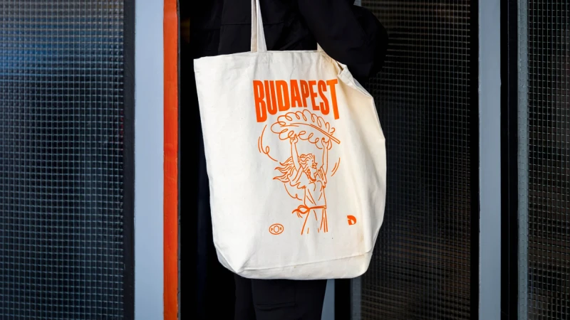 Cotton tote bag with orange Budapest illustration and a woman holding a plant. Worn by a person in a dark coat against a grey tile wall.