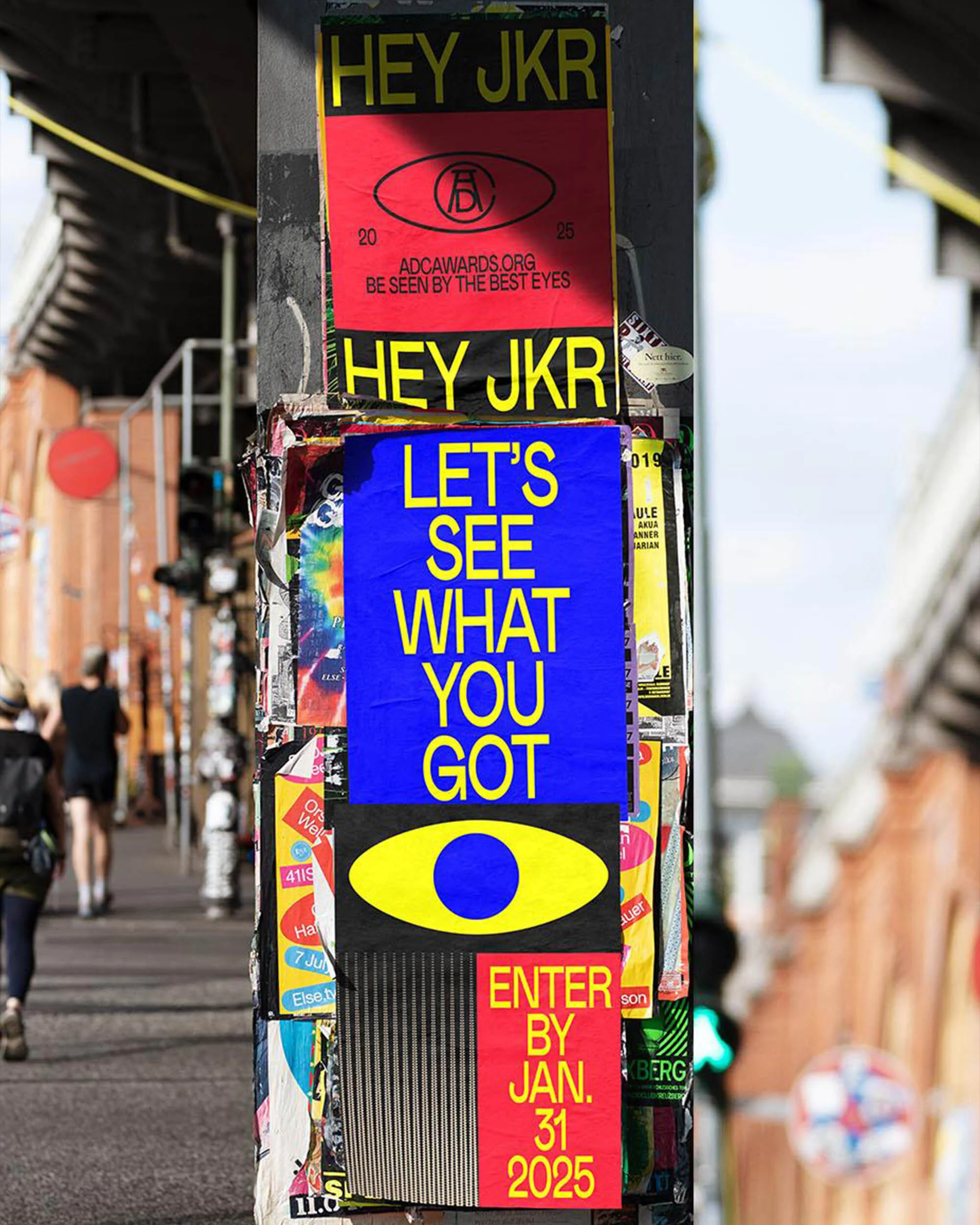 Street art collage of posters on a pole in an urban environment, featuring bold text and graphic elements, with people in background.
