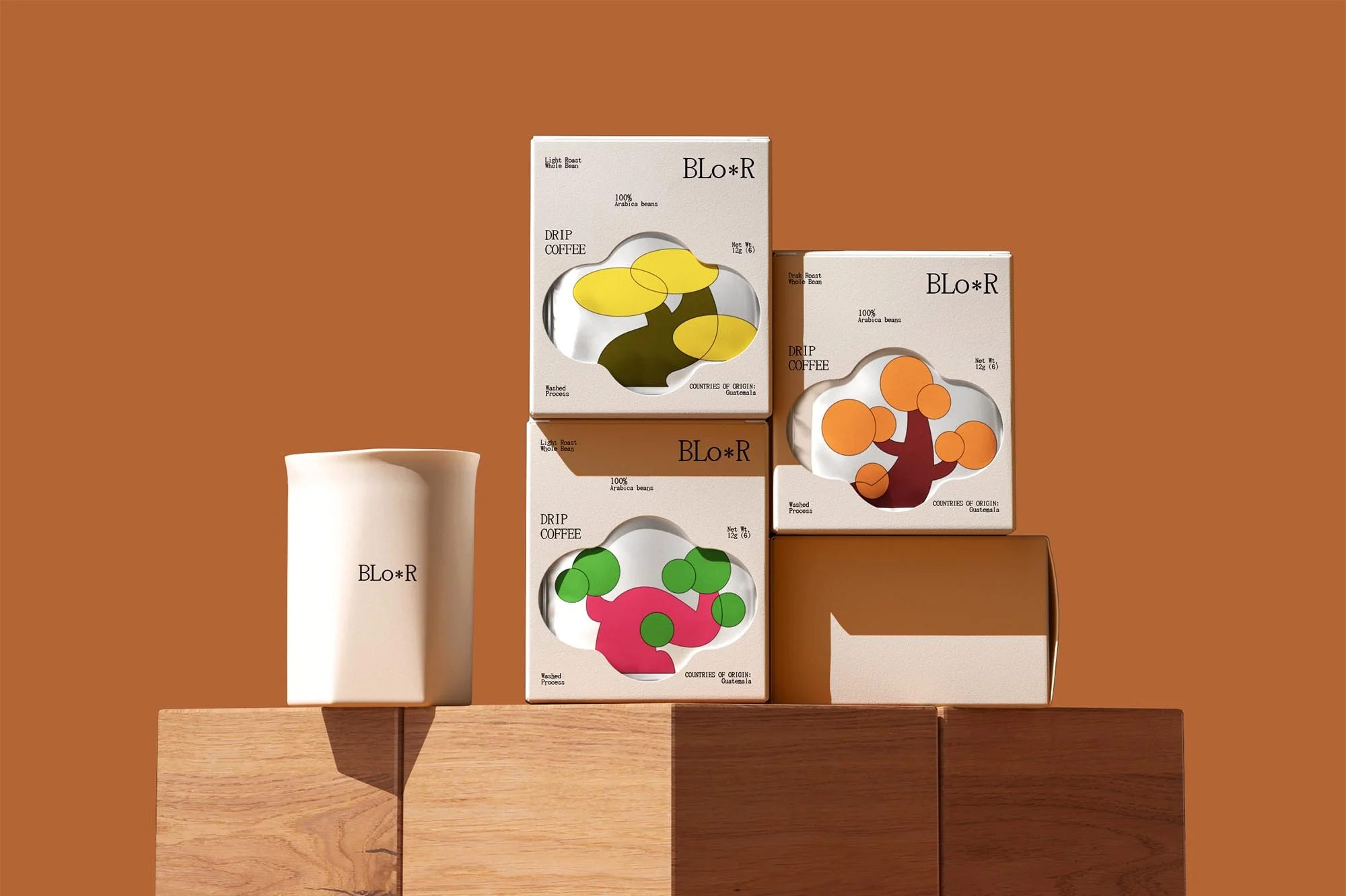 Stacks of BLO*R drip coffee packages on wooden blocks, against a solid brown background, showcasing the brand's packaging design.