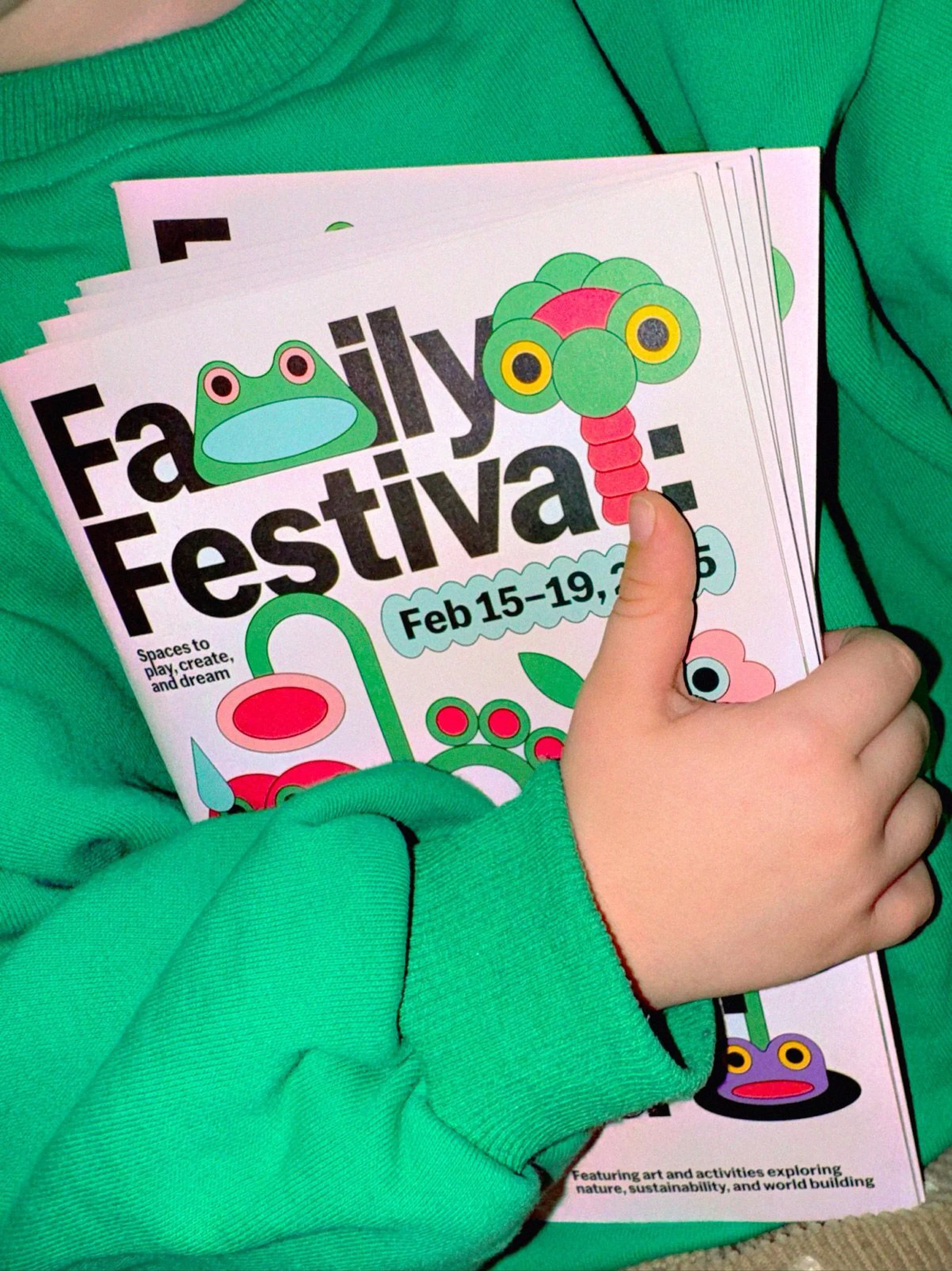 Child in a green sweatshirt holds a stack of Family Festival posters featuring whimsical cartoon frogs, flowers, and palm tree designs.