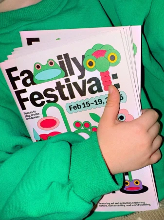 Child in a green sweatshirt holds a stack of Family Festival posters featuring whimsical cartoon frogs, flowers, and palm tree designs.