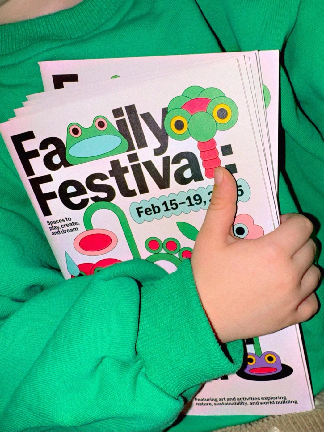 Close-up of hands holding colorful event flyers featuring whimsical cartoon illustrations against a bright green sweatshirt.