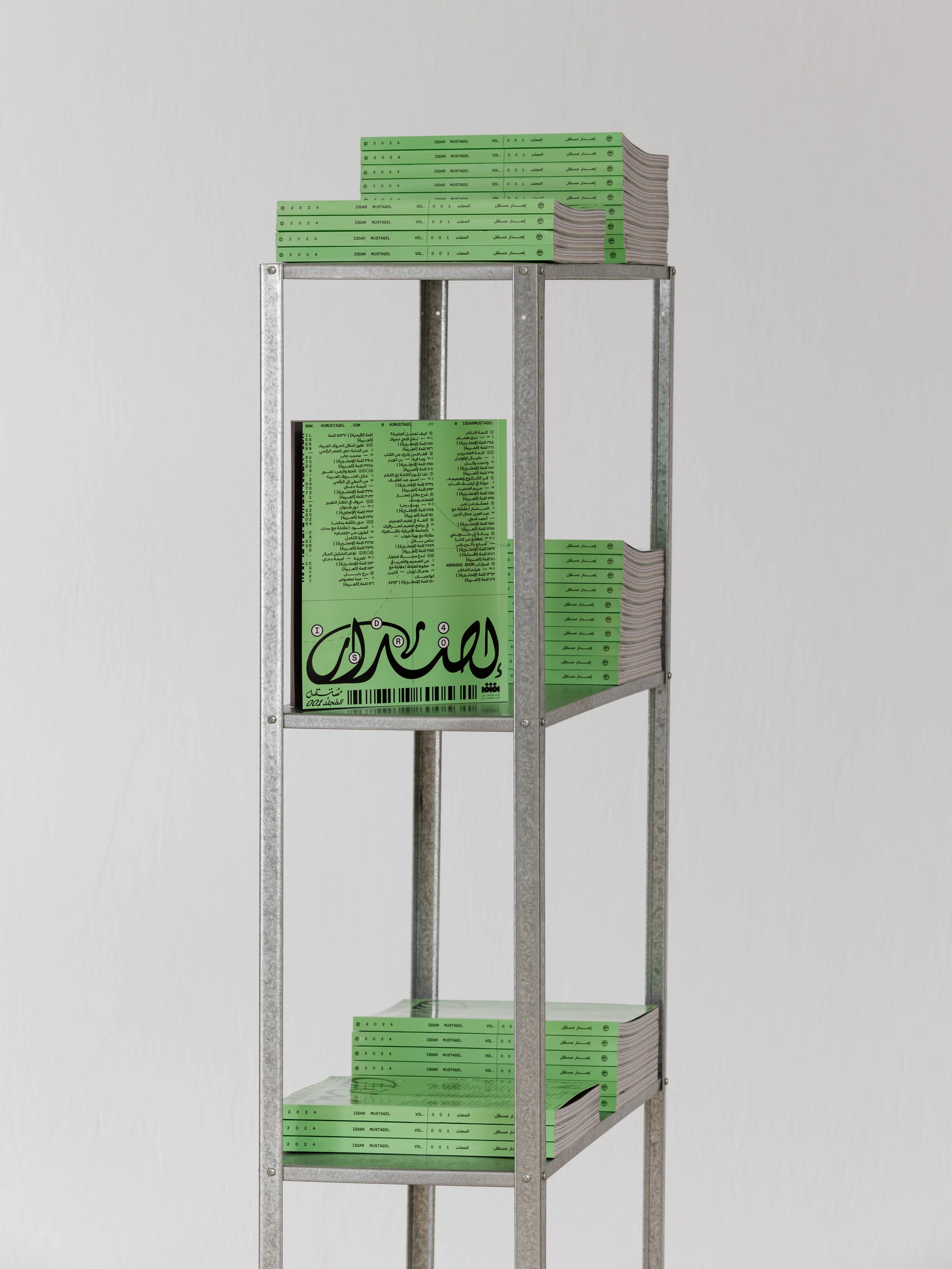 Silver metal shelving unit with stacks of light green magazines with black text on a light gray background in a neutral-lit studio.