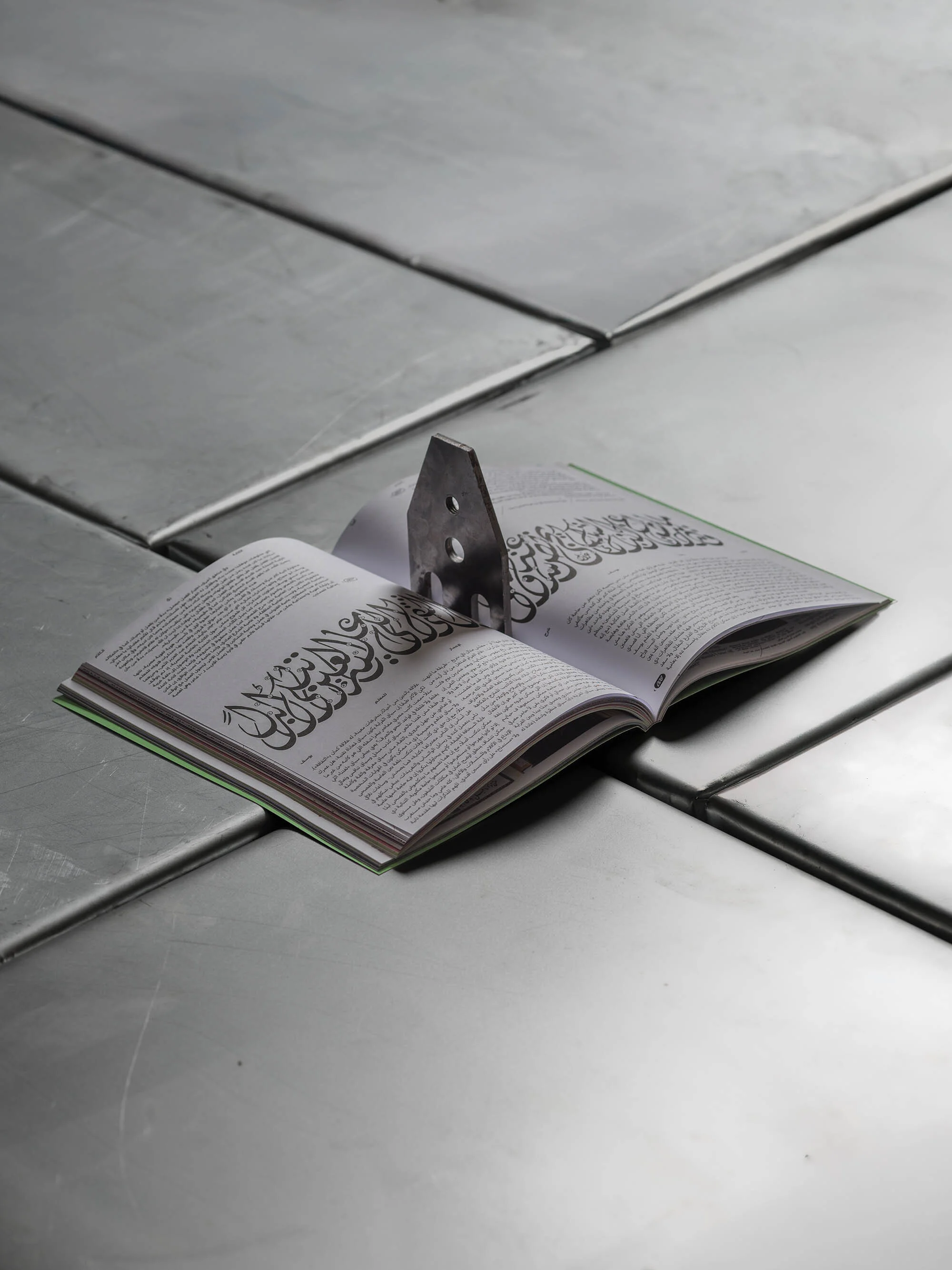 Open book with Arabic script held open by a metal bookmark on a tiled metal surface, creating a minimalist and contemplative image.