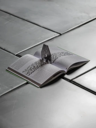 Open book with Arabic script held open by a metal bookmark on a tiled metal surface, creating a minimalist and contemplative image.