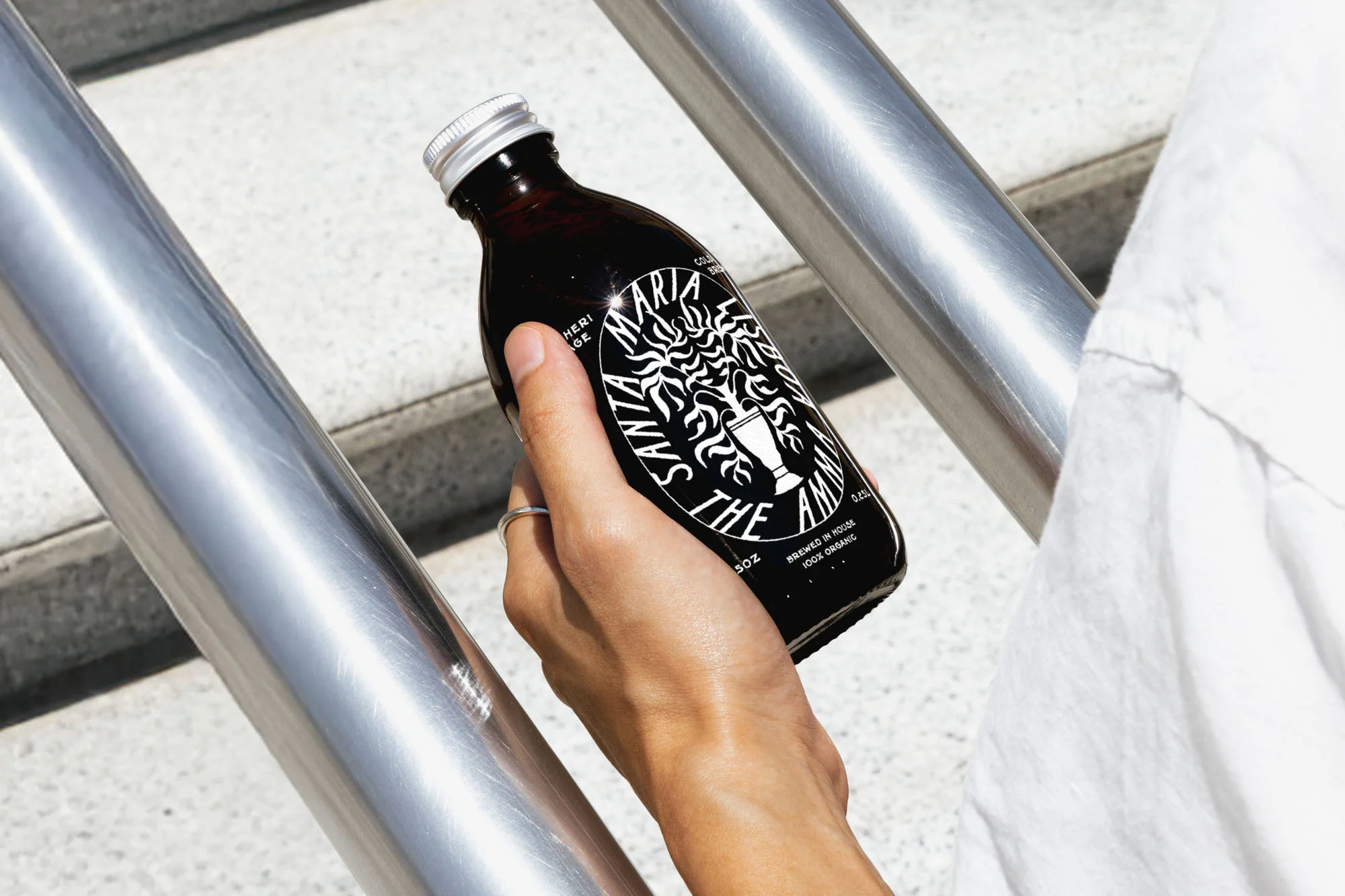 A person holds a dark-colored glass bottle of Maria Santa The Amoroso organic beverage in front of a metal railing outdoors.