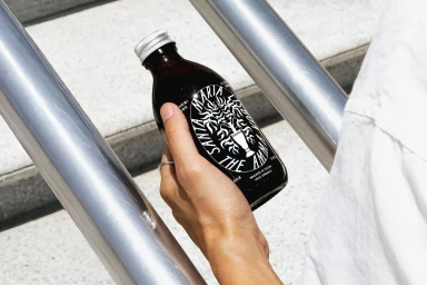 Hand holding a dark glass bottle with custom typography label outdoors near metal railings