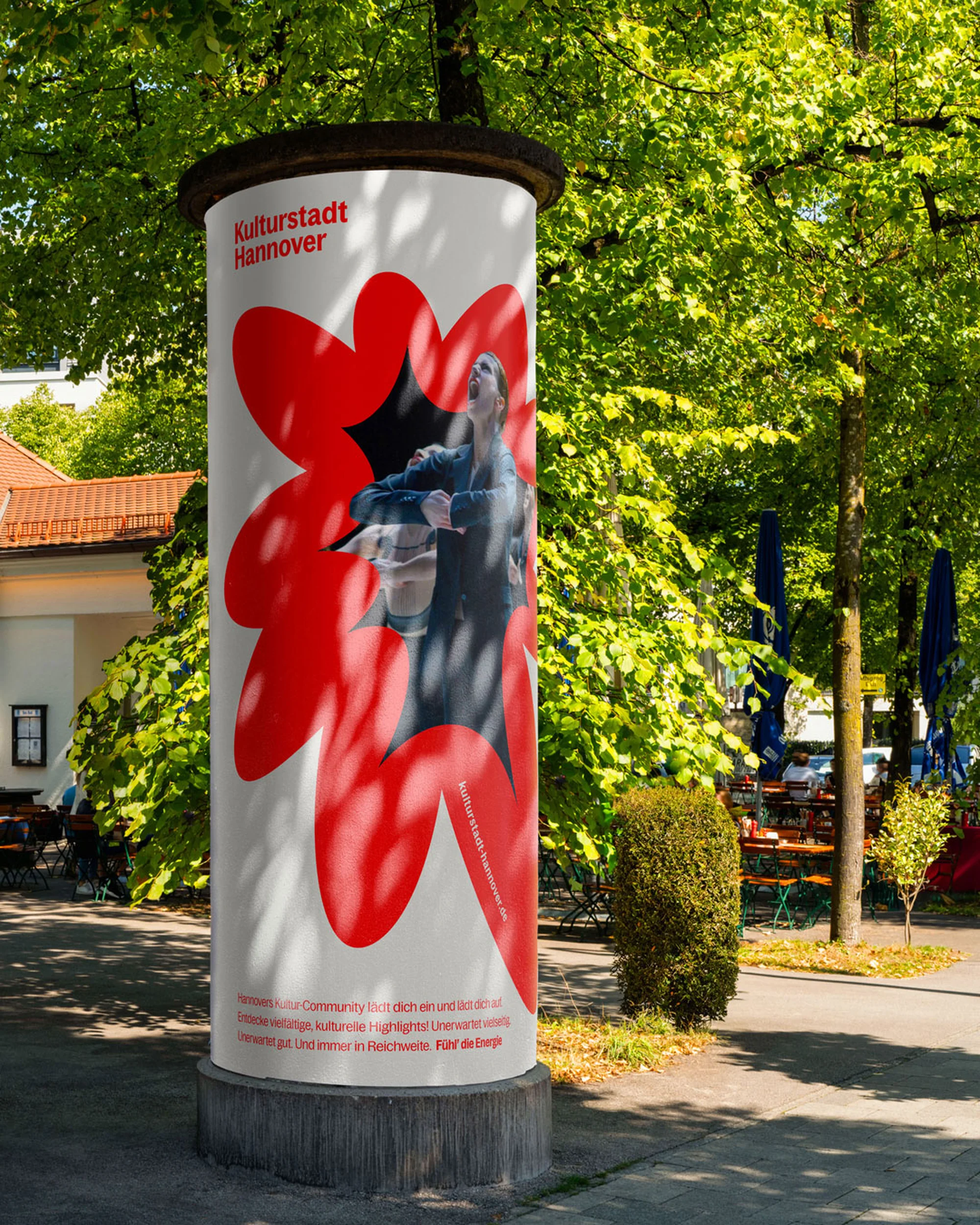 Outdoor shot of a cylindrical advertising column with the words 'Kulturstadt Hannover' in front of a bright green tree canopy.