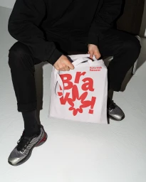 Person seated on a chair holding a white tote bag with red typographic branding against a white seamless background.
