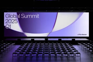 Empty auditorium with neat rows of dark chairs facing a wide presentation screen displaying purple abstract graphics and text.