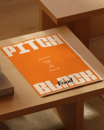 Orange flyer with white block lettering resting on a light wooden table inside a minimal room.