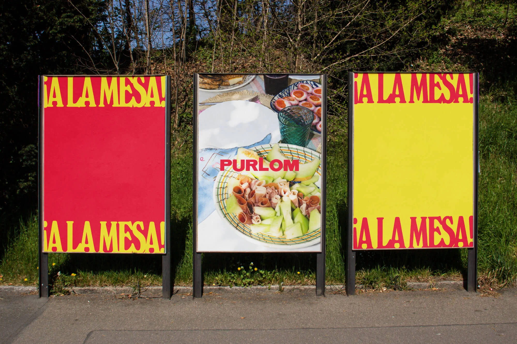 Three colorful advertising billboards featuring the text '¡A LA MESA!' and food imagery against a natural outdoor backdrop.