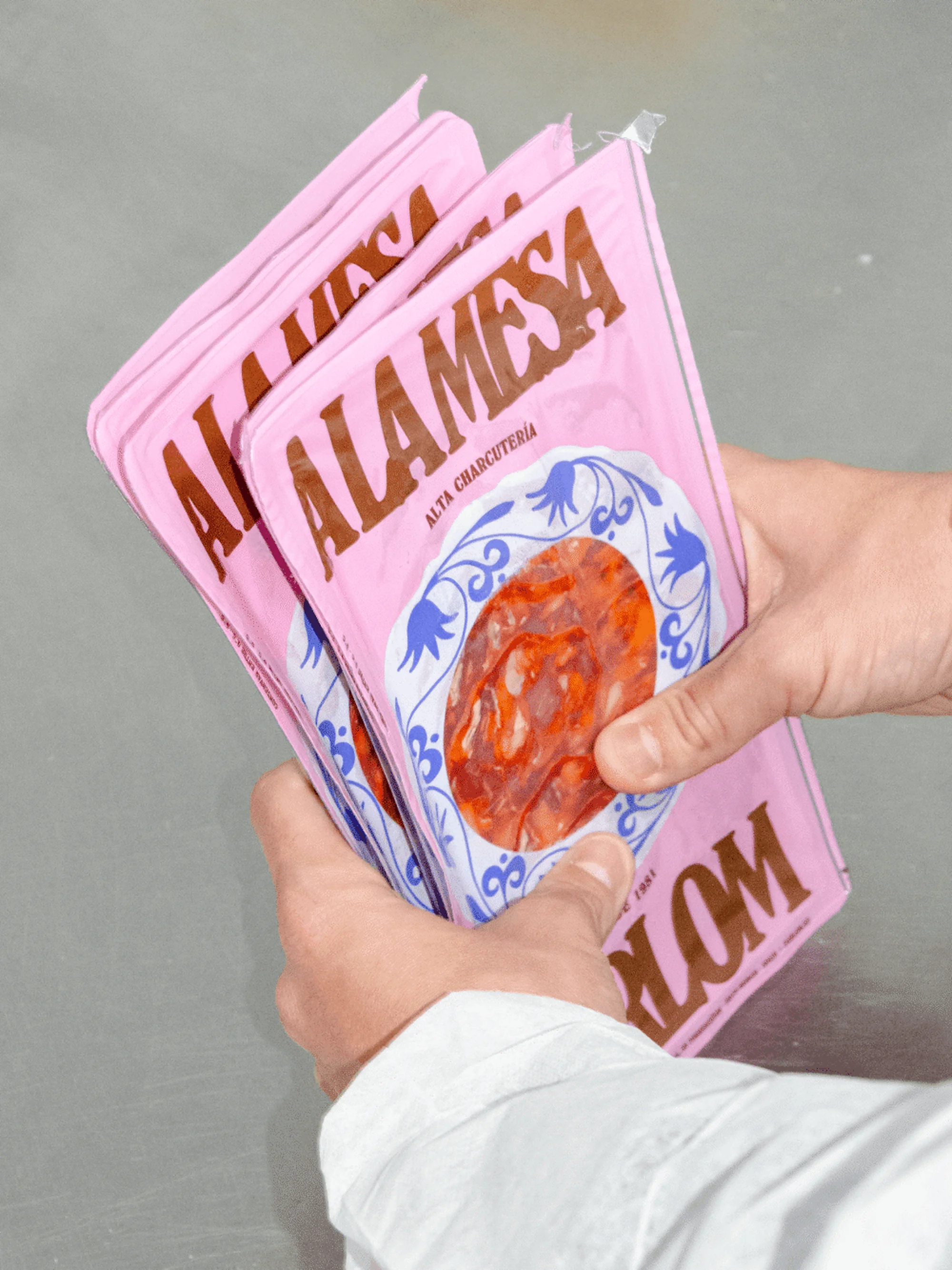 Close-up of hands holding pink packages of Alamesa charcuterie, featuring clear windows revealing the cured meat inside.