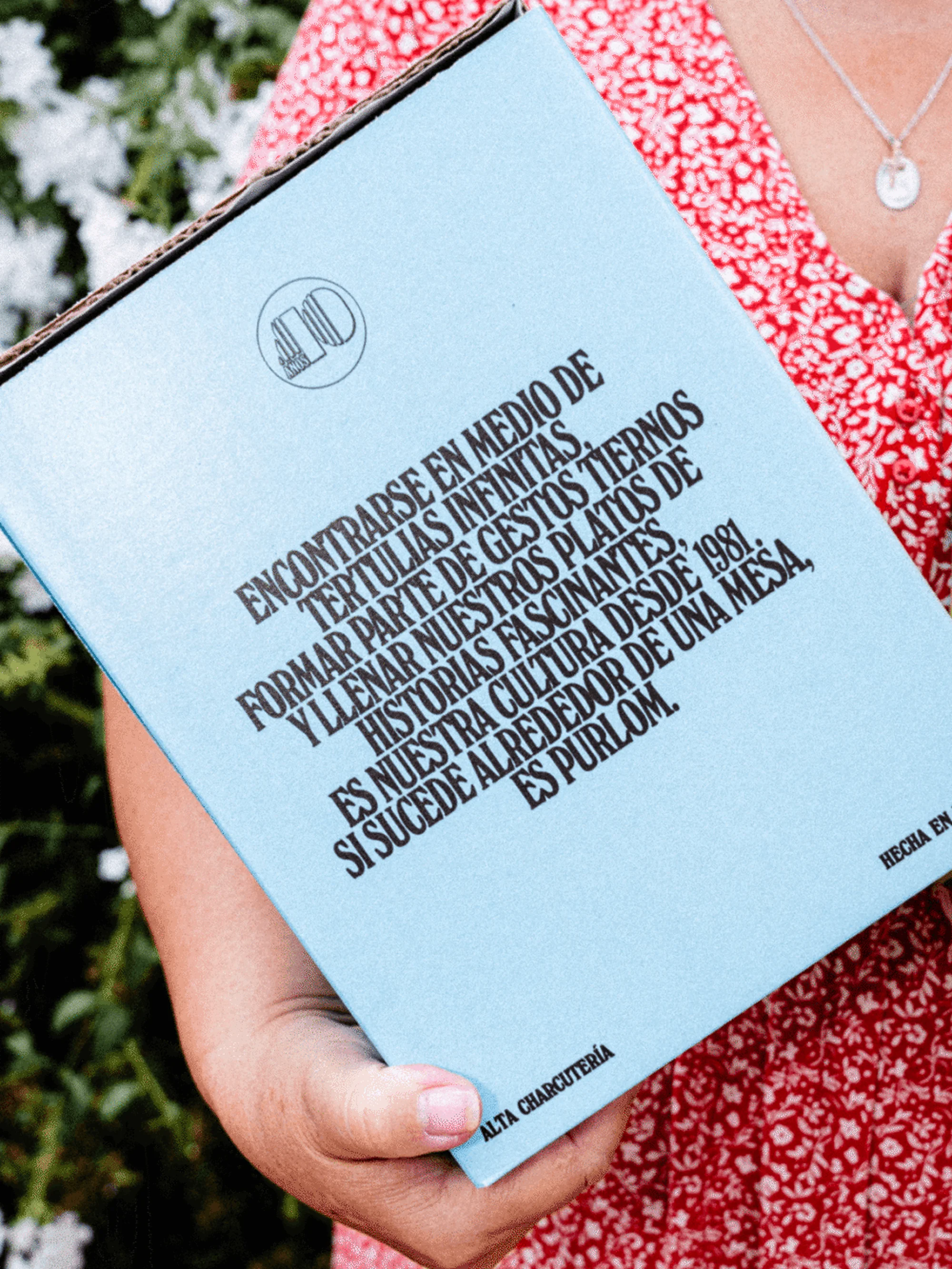 Close-up of person holding a light blue box with black text, featuring food-related marketing copy and a logo. Red floral dress visible.