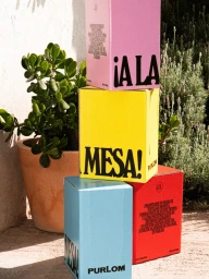Stacked colorful packaging boxes in pink, yellow, blue, and red with bold typography next to potted green plants outdoors.