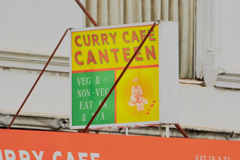 Bright, graphic sign for 'Curry Cafe Canteen' with vegetarian options, featuring orange, yellow, and green colors and sun illustration.