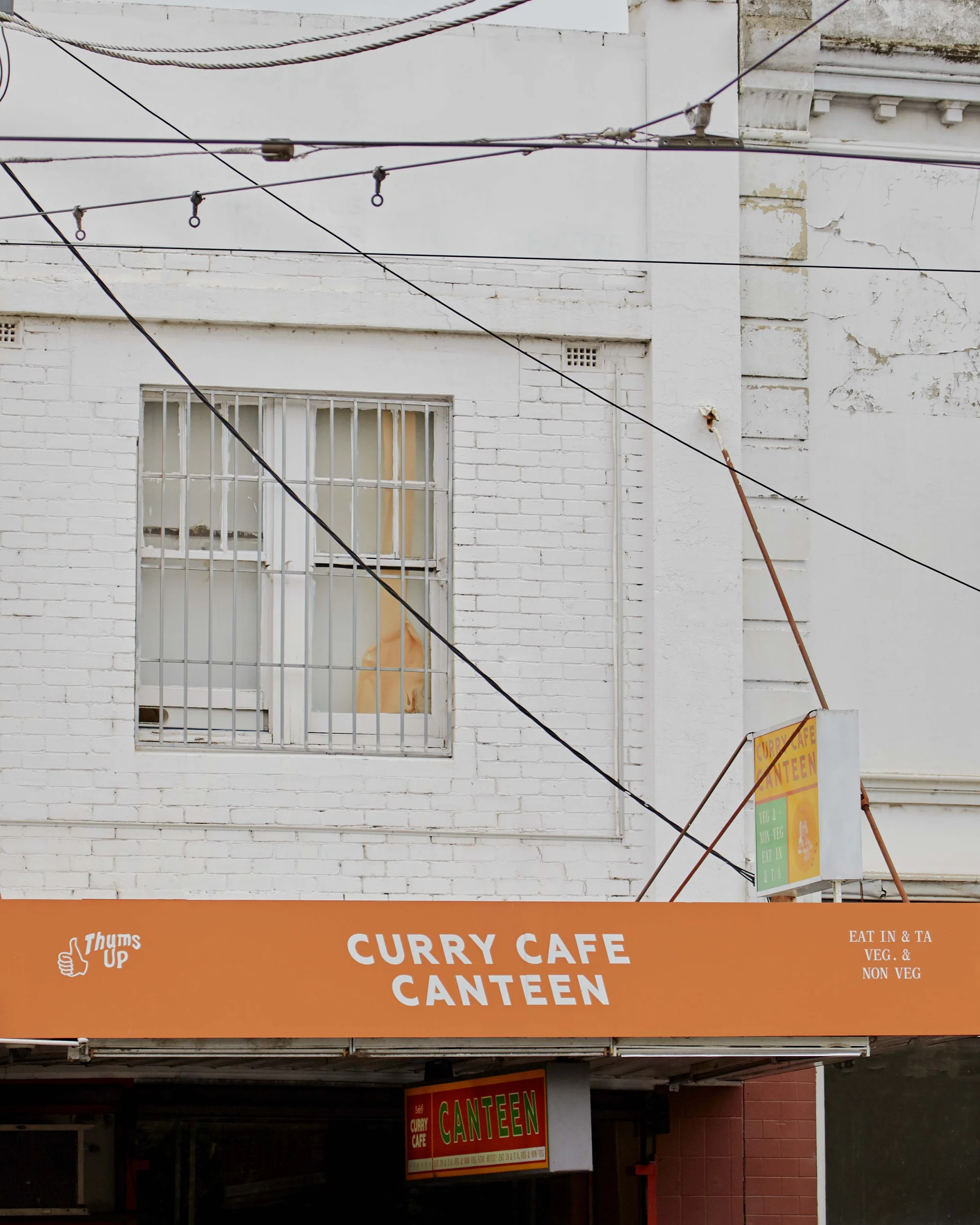 Exterior of Curry Cafe Canteen with bright orange signage, window with bars, and overhead wires against a white brick building.