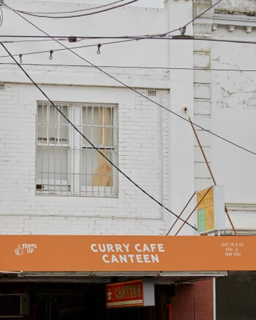 Exterior of Curry Cafe Canteen with bright orange signage, window with bars, and overhead wires against a white brick building.